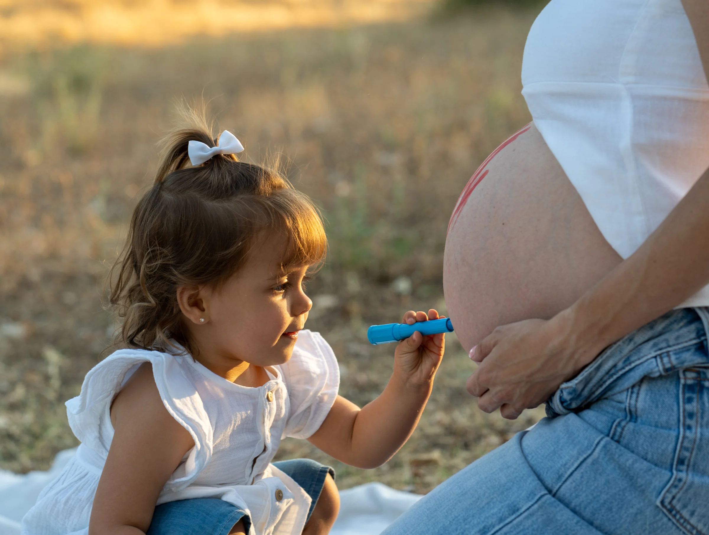 Niña pinta con un rotulador azul sobre la barriga de su madre embarazada mientras la madre la observa.