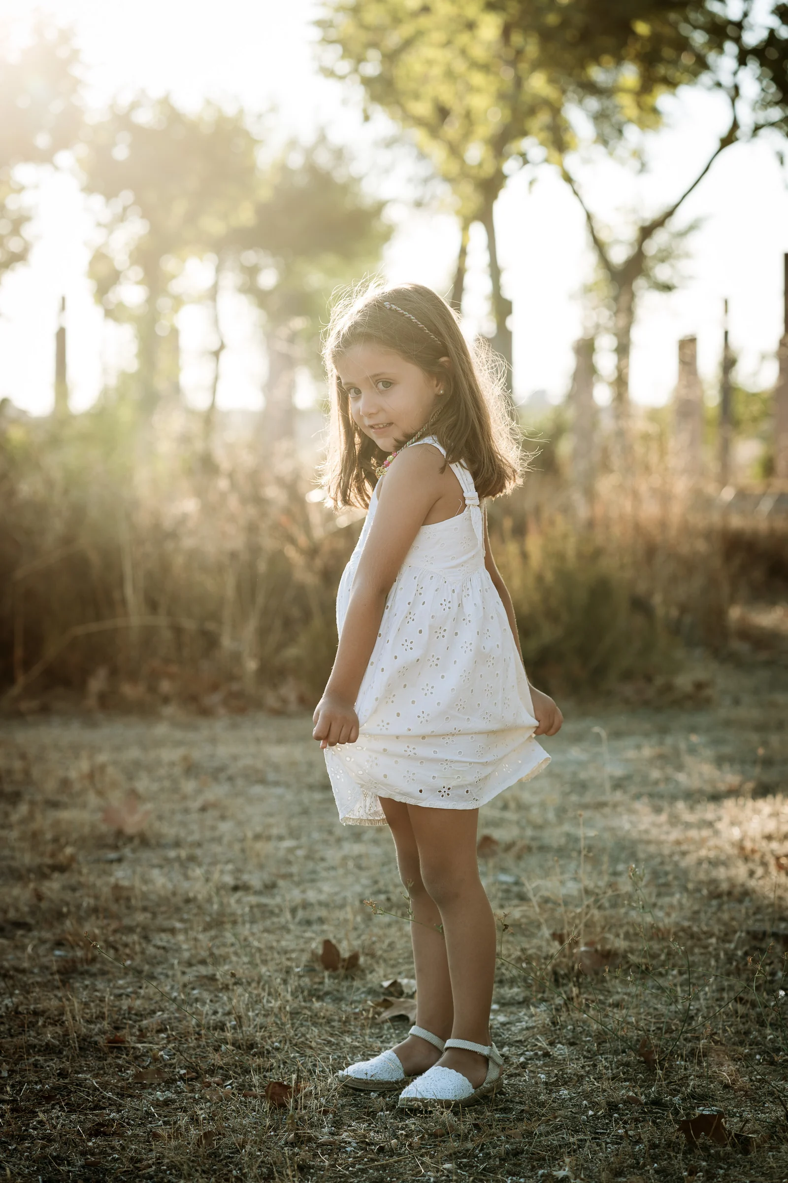 Niña con vestido blanco posa de pie en un parque durante una sesión familiar al atardecer.