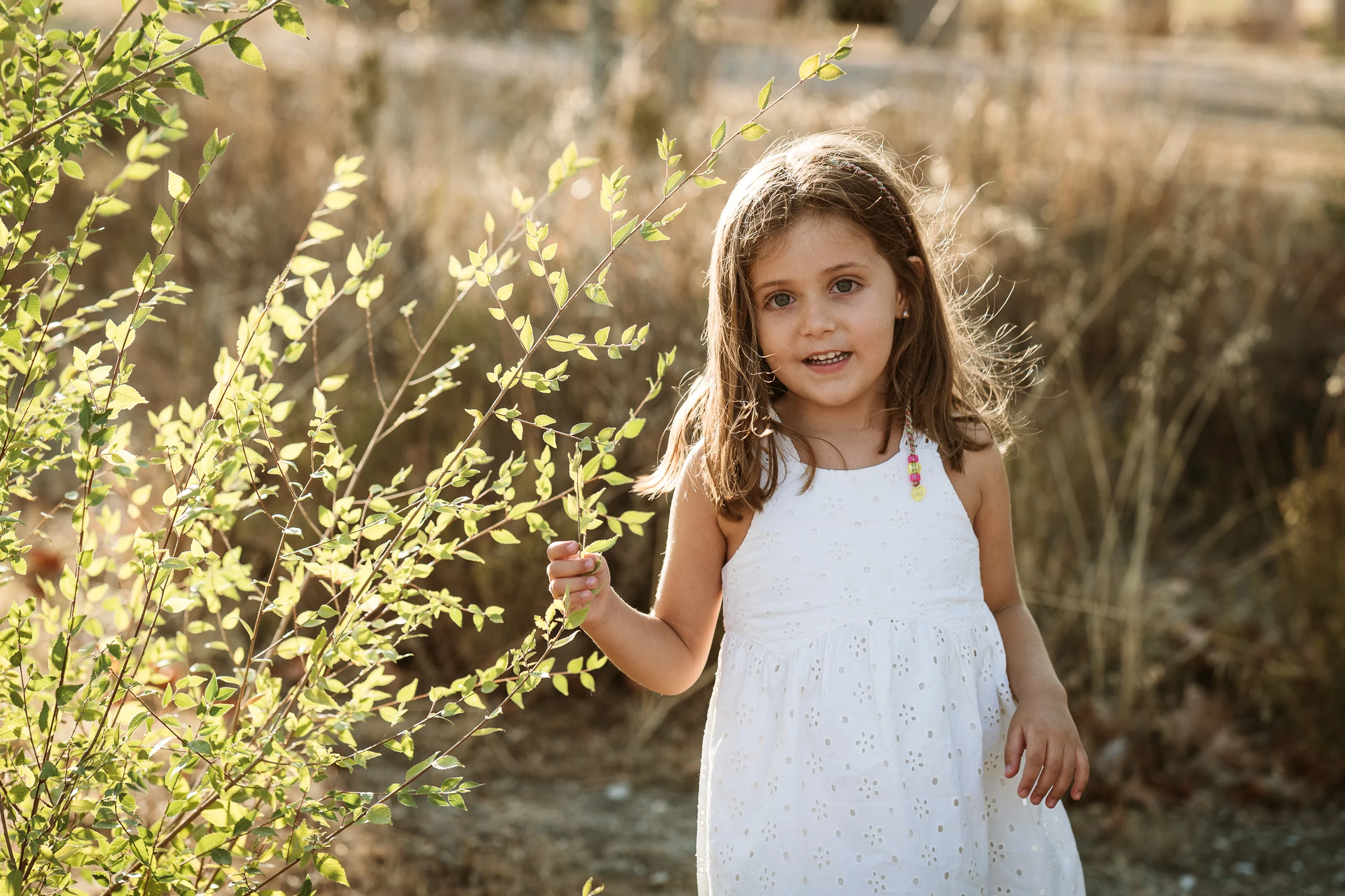 Niña con vestido blanco mira a cámara junto a unas ramas iluminadas por la luz del atardecer.