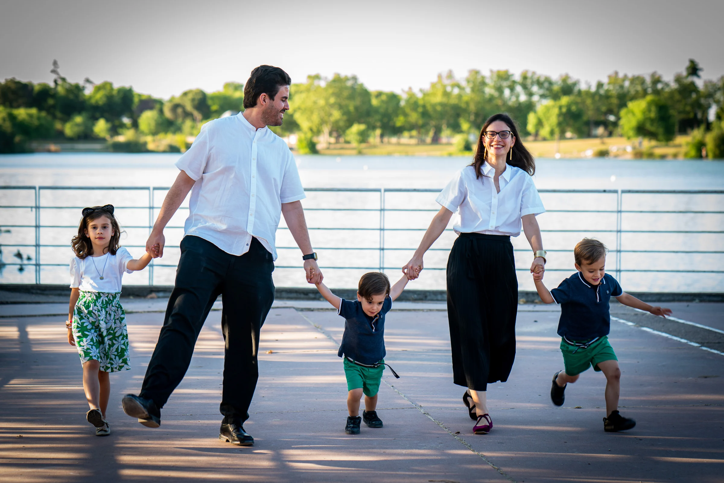 Familia de cinco personas camina de la mano junto a un lago durante una sesión fotográfica.