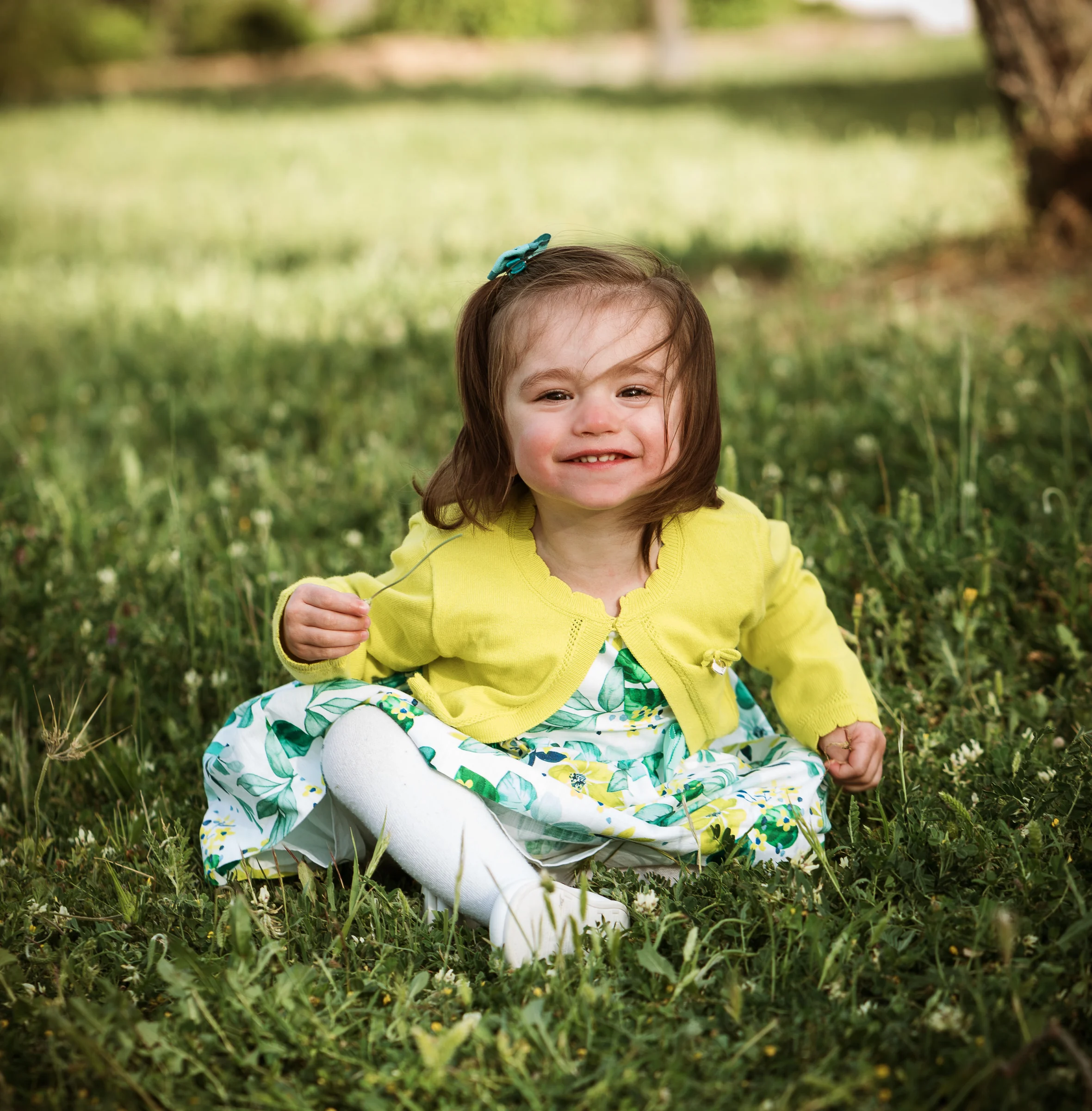 Niña pequeña con vestido floral verde y chaqueta amarilla sonríe sentada sobre el césped.