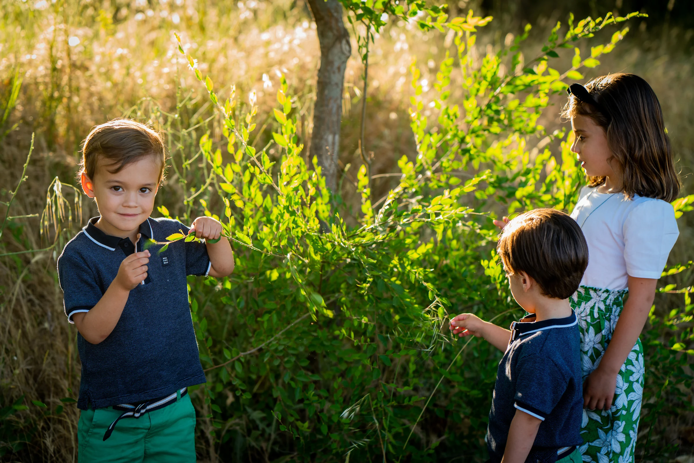 Tres hermanos juegan con unas ramas durante una sesión familiar al aire libre.