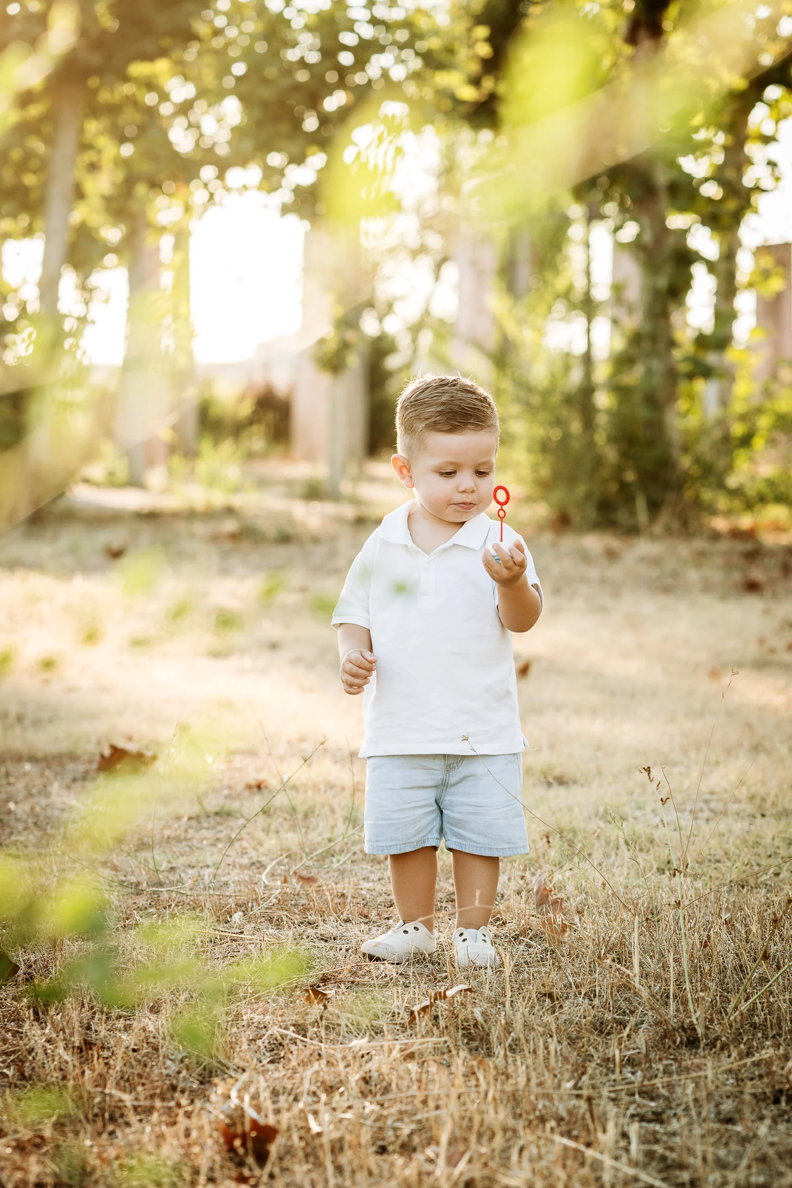 Niño pequeño vestido de blanco observa un juguete de burbujas en un entorno natural al atardecer.
