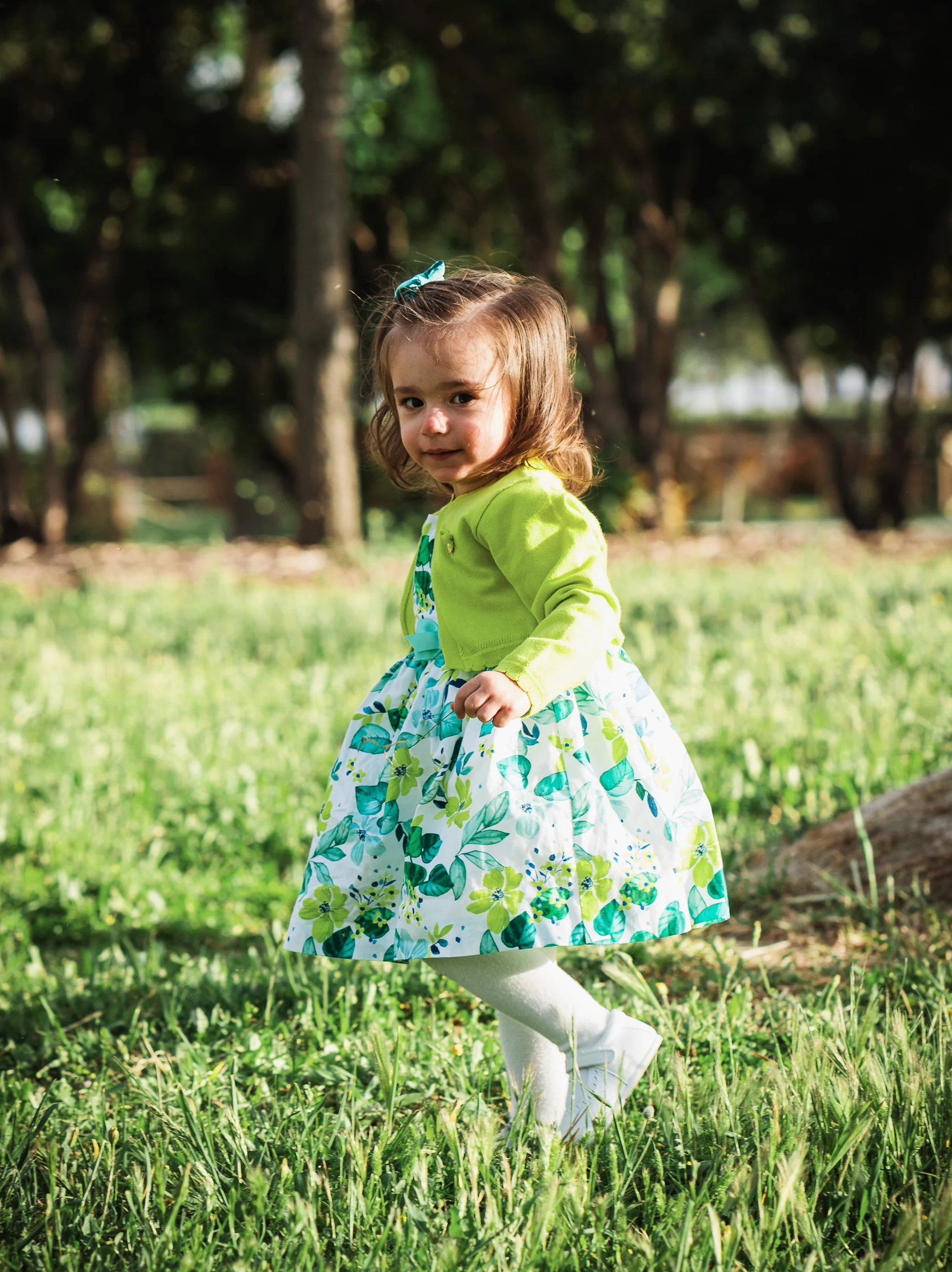 Niña pequeña con vestido floral camina entre la hierba durante una sesión infantil al aire libre.