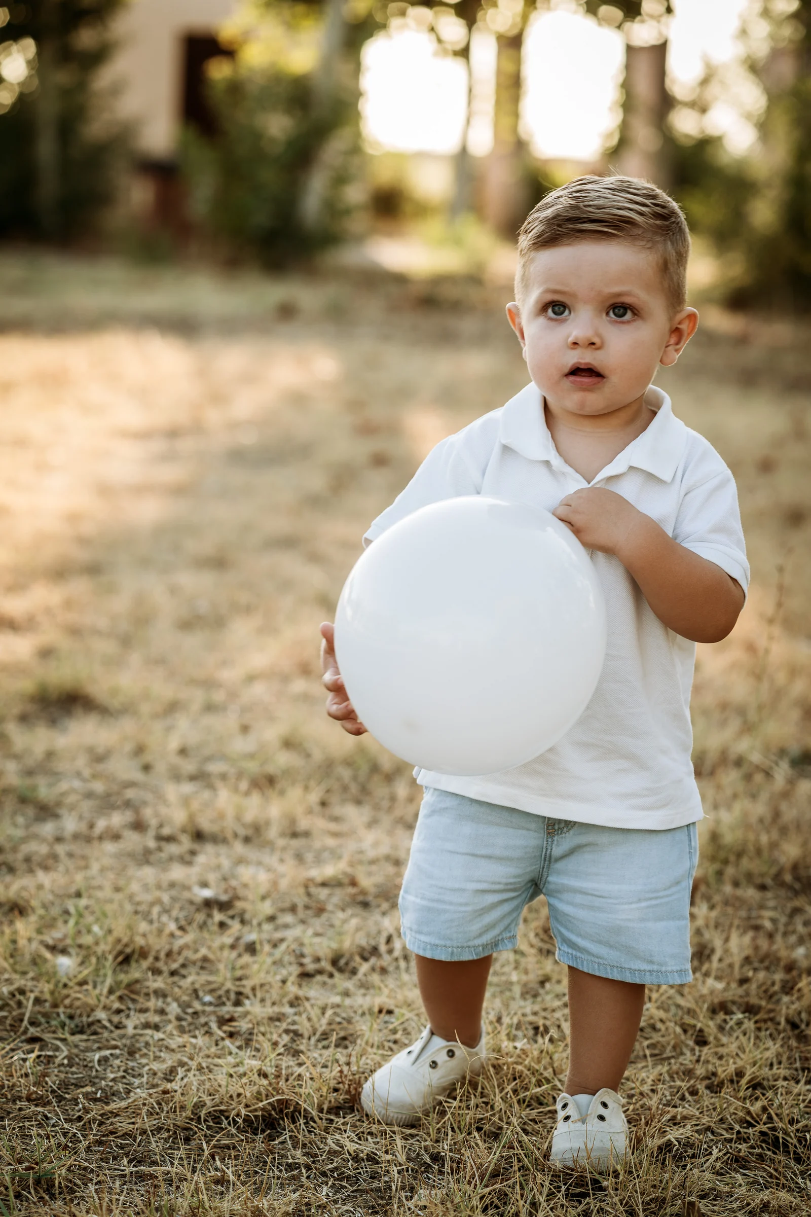 Niño pequeño con polo blanco sostiene un globo blanco en un campo de hierba seca.