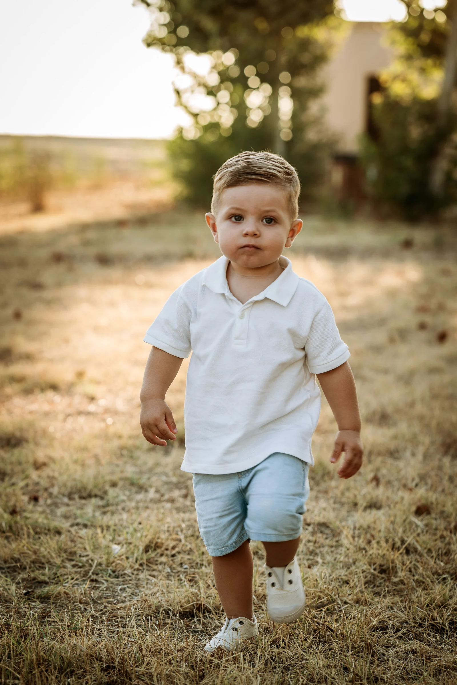 Niño pequeño vestido de blanco camina hacia la cámara por un campo iluminado al atardecer.