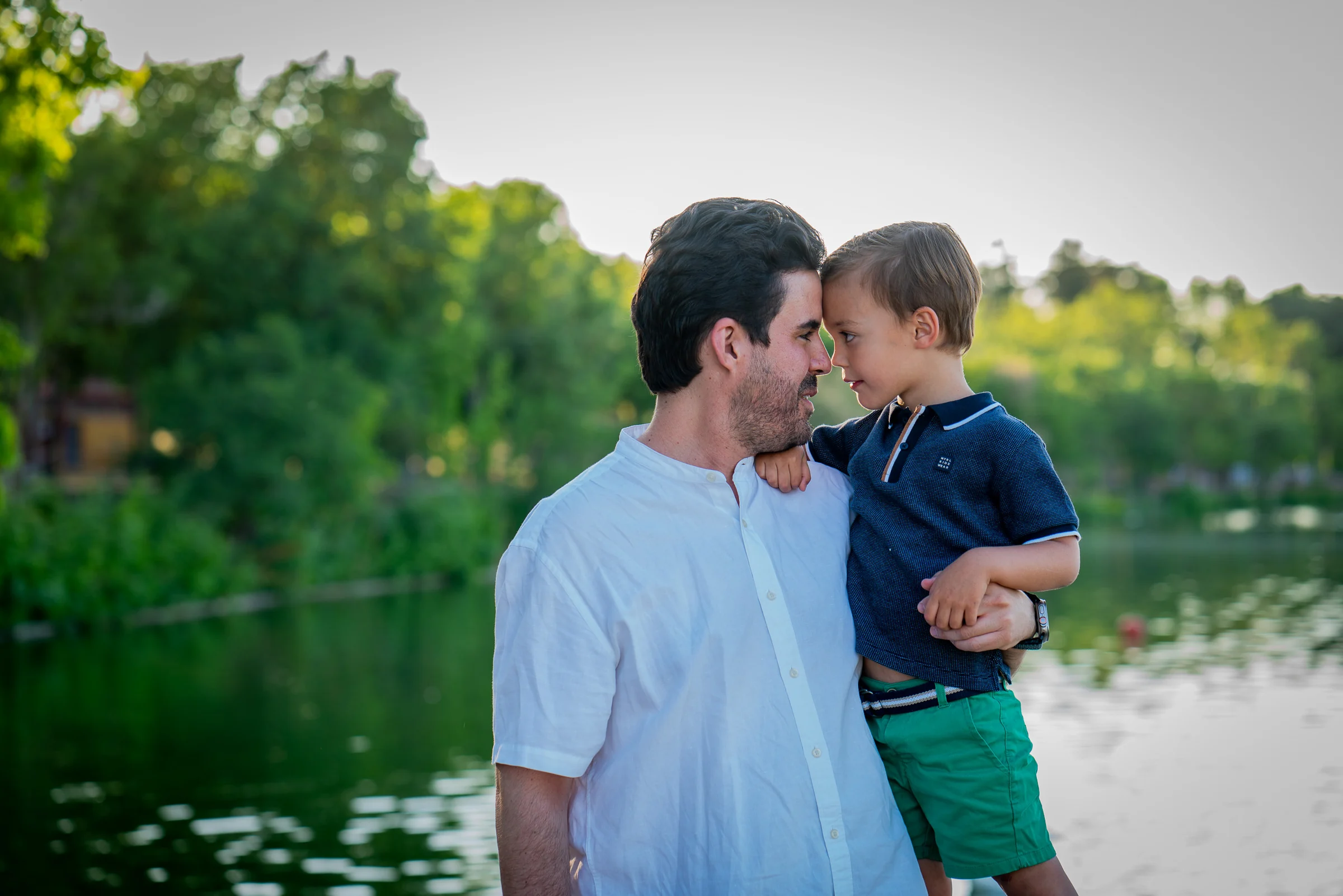Padre sostiene a su hijo pequeño y ambos juntan sus frentes junto a un lago.