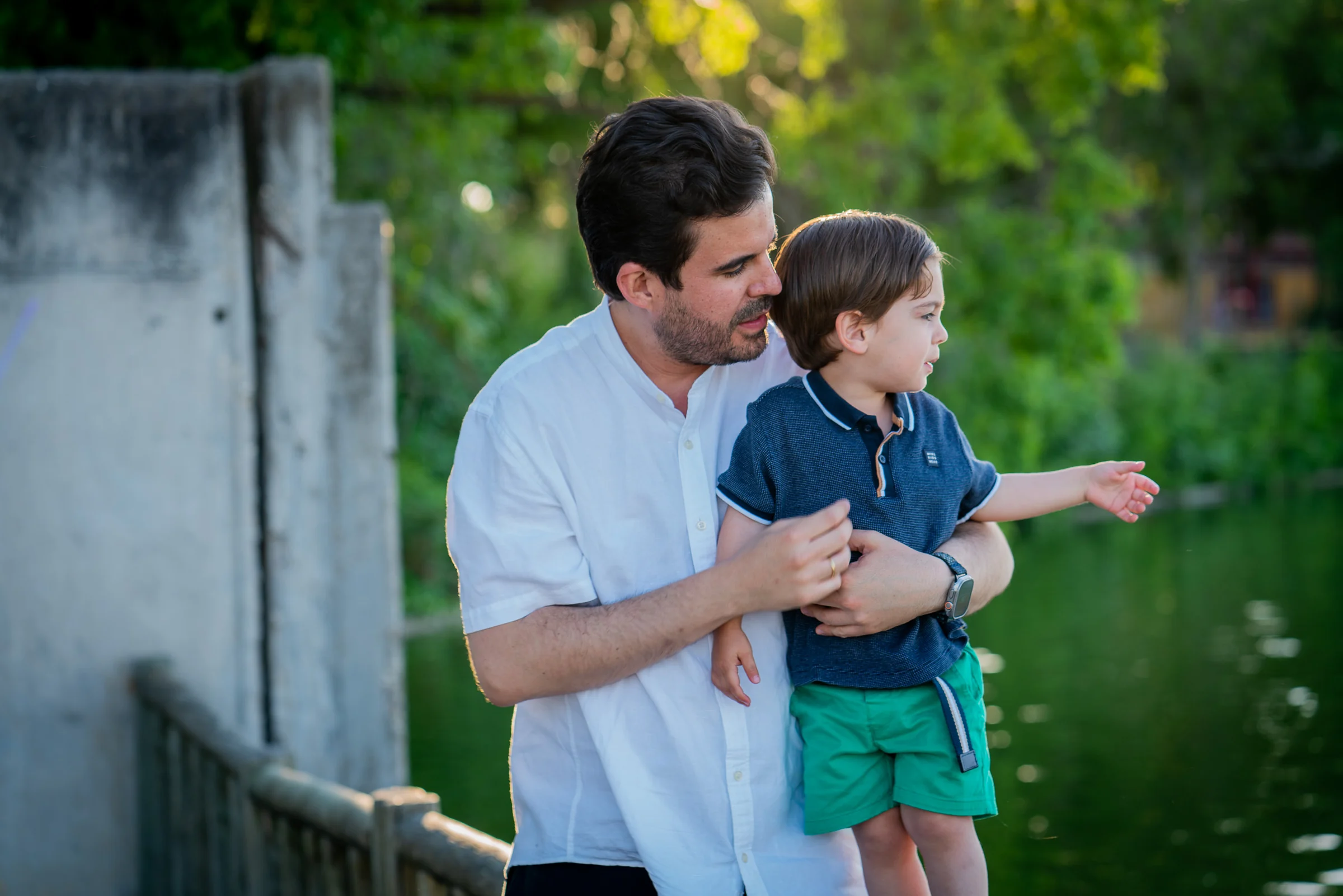 Padre sostiene a su hijo pequeño mientras el niño señala algo junto a un lago.
