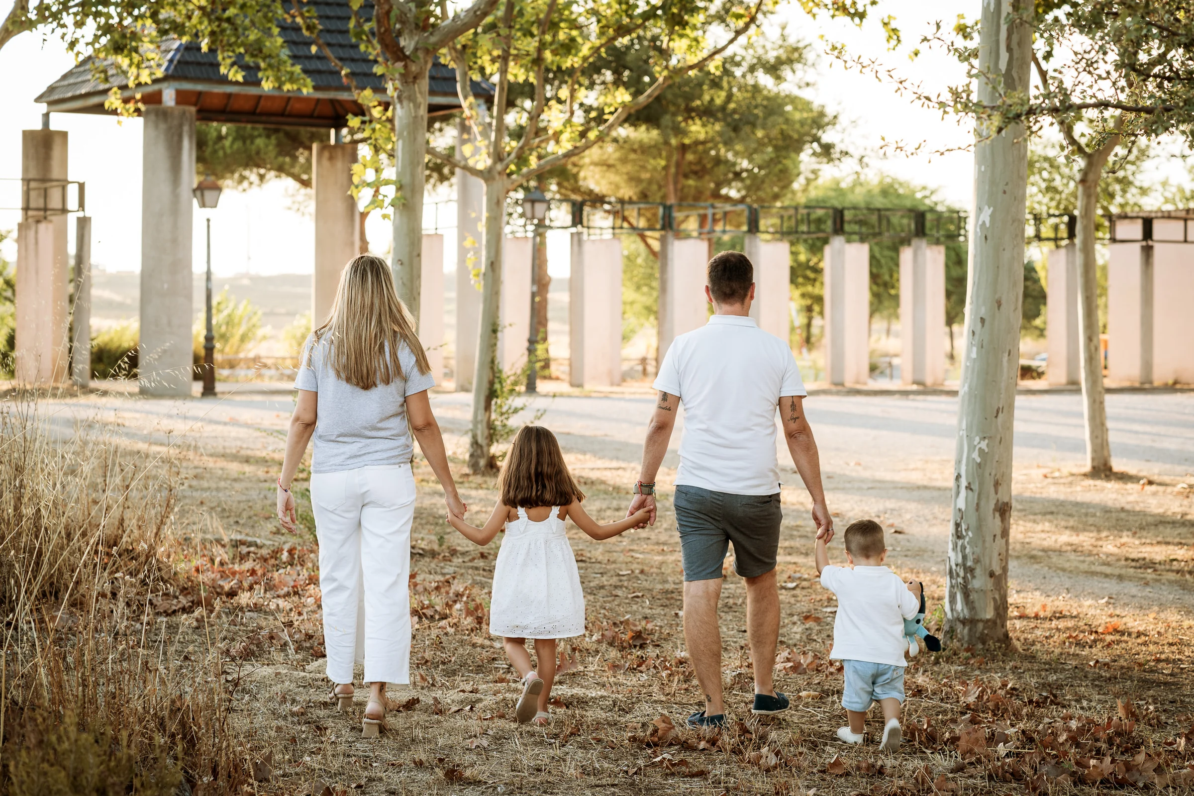 Familia de cuatro camina de espaldas cogida de la mano por un parque.