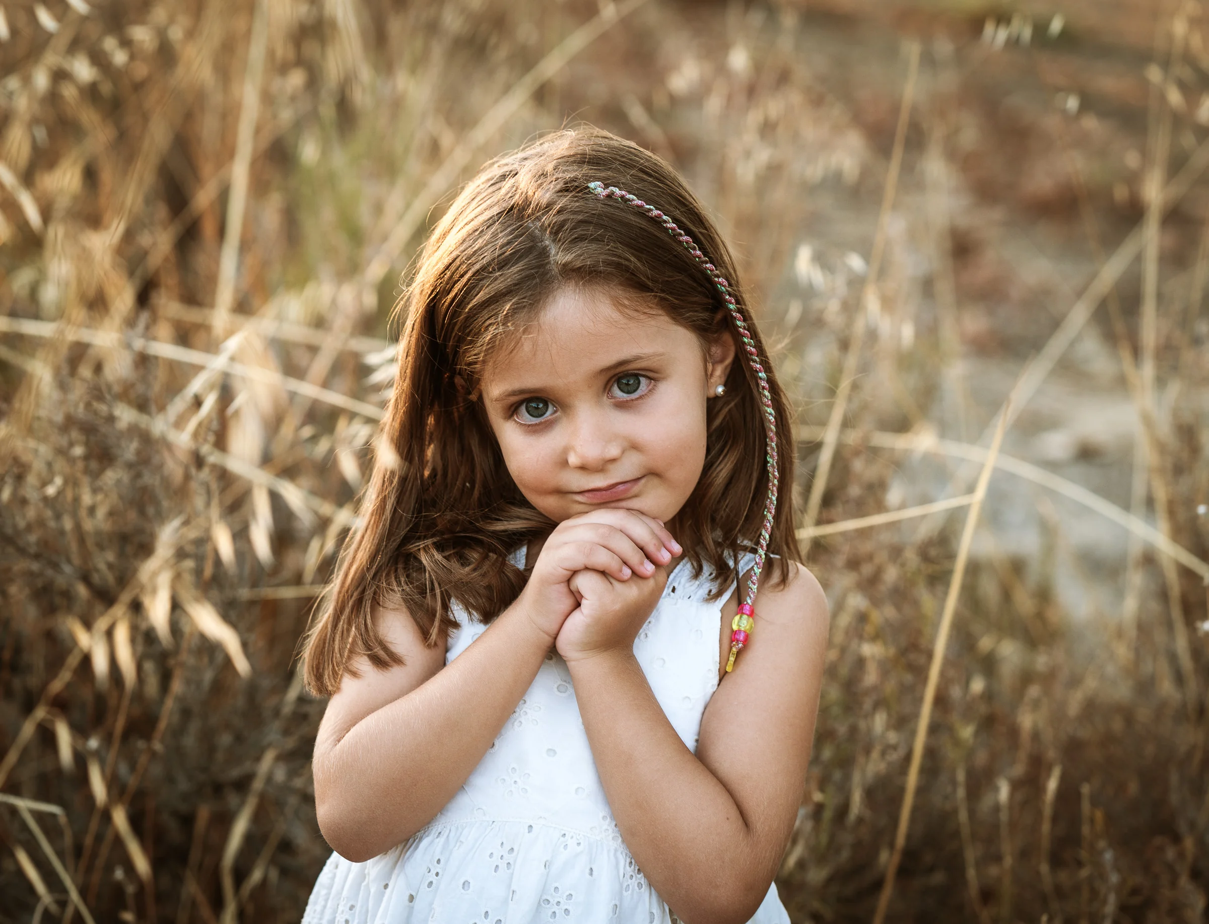 Niña con vestido blanco posa con las manos bajo la barbilla entre hierba seca.