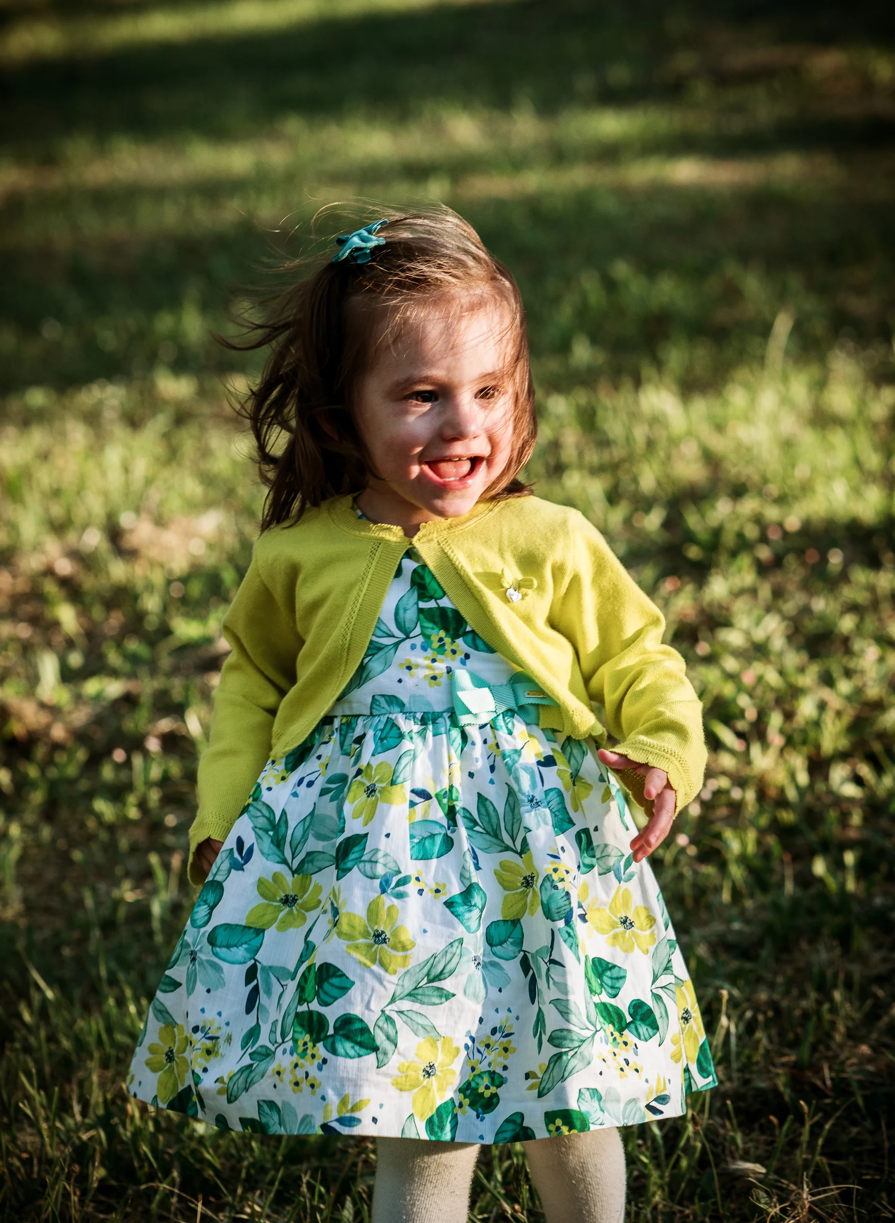 Niña pequeña con vestido floral y chaqueta amarilla sonríe de pie sobre la hierba.