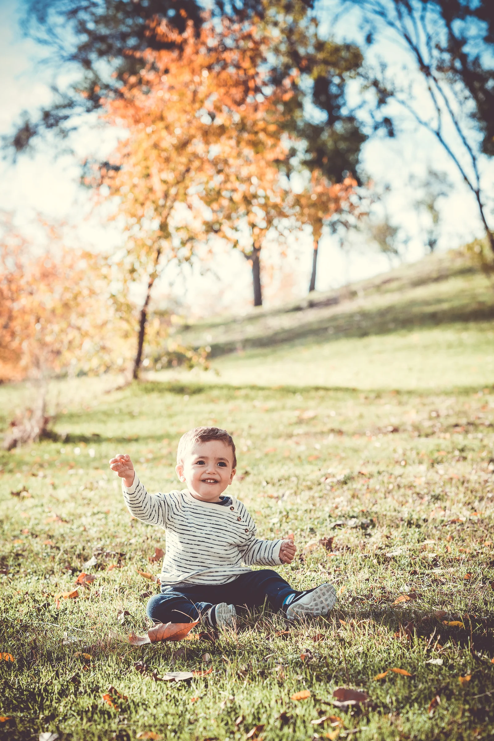 Niño pequeño sonríe sentado sobre el césped rodeado de hojas de otoño.