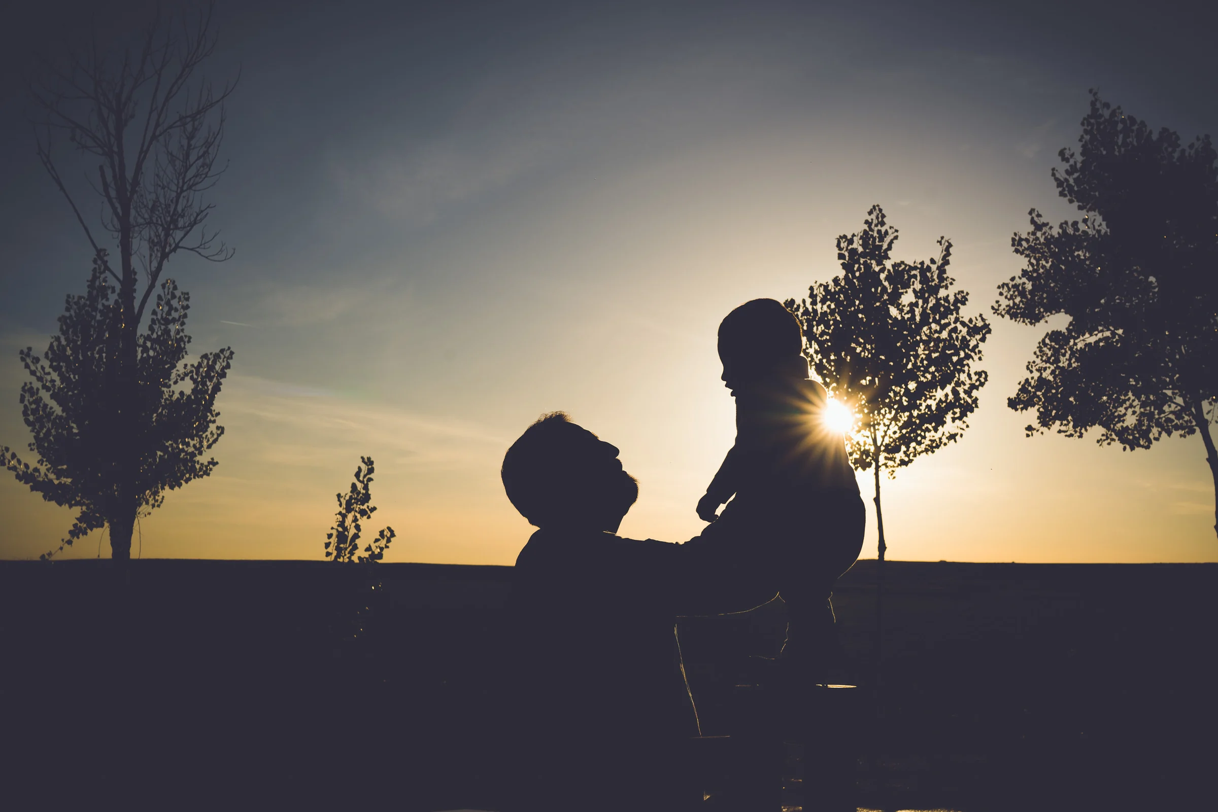 Silueta de un padre sosteniendo a su hijo pequeño frente al sol del atardecer.