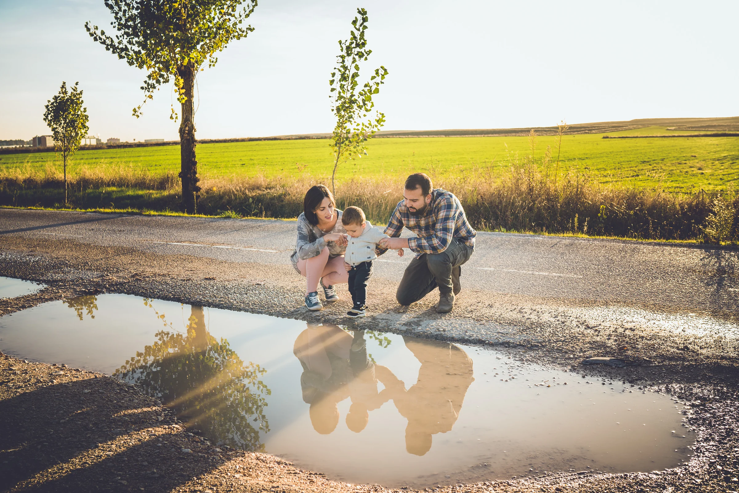 Familia de tres se agacha junto a un charco donde se reflejan sus figuras y los árboles del paisaje.
