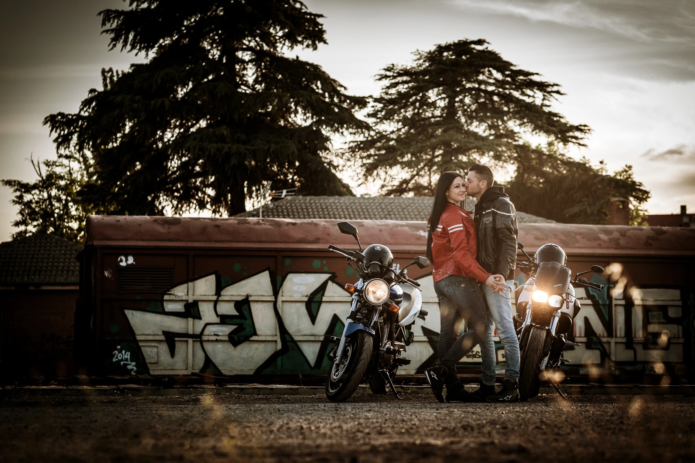 Sara y Edu se besan entre sus motos al atardecer en un entorno ferroviario de Aranjuez durante su preboda.
